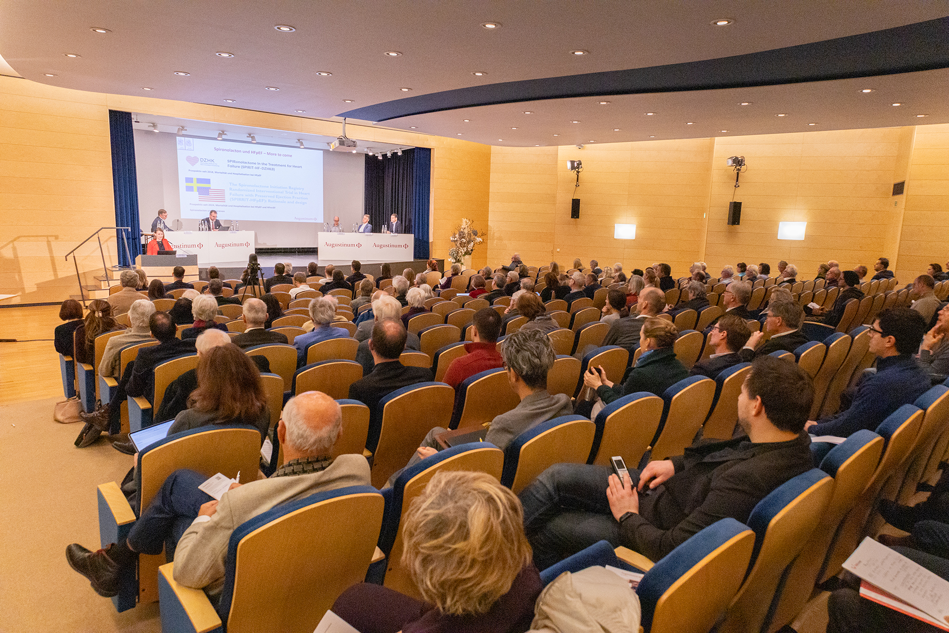 Augustinum Klinik Symposium Viele Besucher*innen sitzen in einem Theatersaal und blicken auf die Veranstalter, die auf der Bühne sitzen