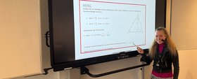 Augustinum Samuel-Heinicke-Realschule Panel Eine Lehrerin, die ein Headset trägt, zeigt lächelnd auf den Bildschirm neben ihr, auf dem Lerninhalte angezeigt werden