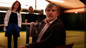 Extrawurst Drei Personen in einer Tennishalle neben einem Tennisnetz; eine Person sitzt im Vordergrund auf einem Stuhl, zwei weitere stehen im Hintergrund auf dem Spielfeld.