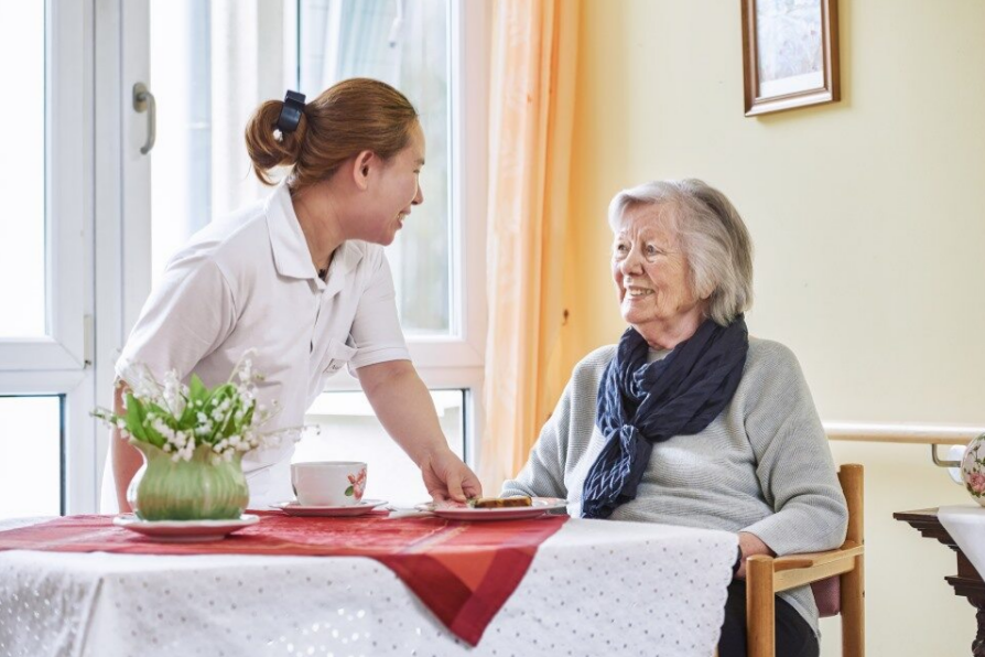 Eine Seniorin sitzt an einem Tisch und eine Mitarbeiterin bringt ihr etwas zu essen 