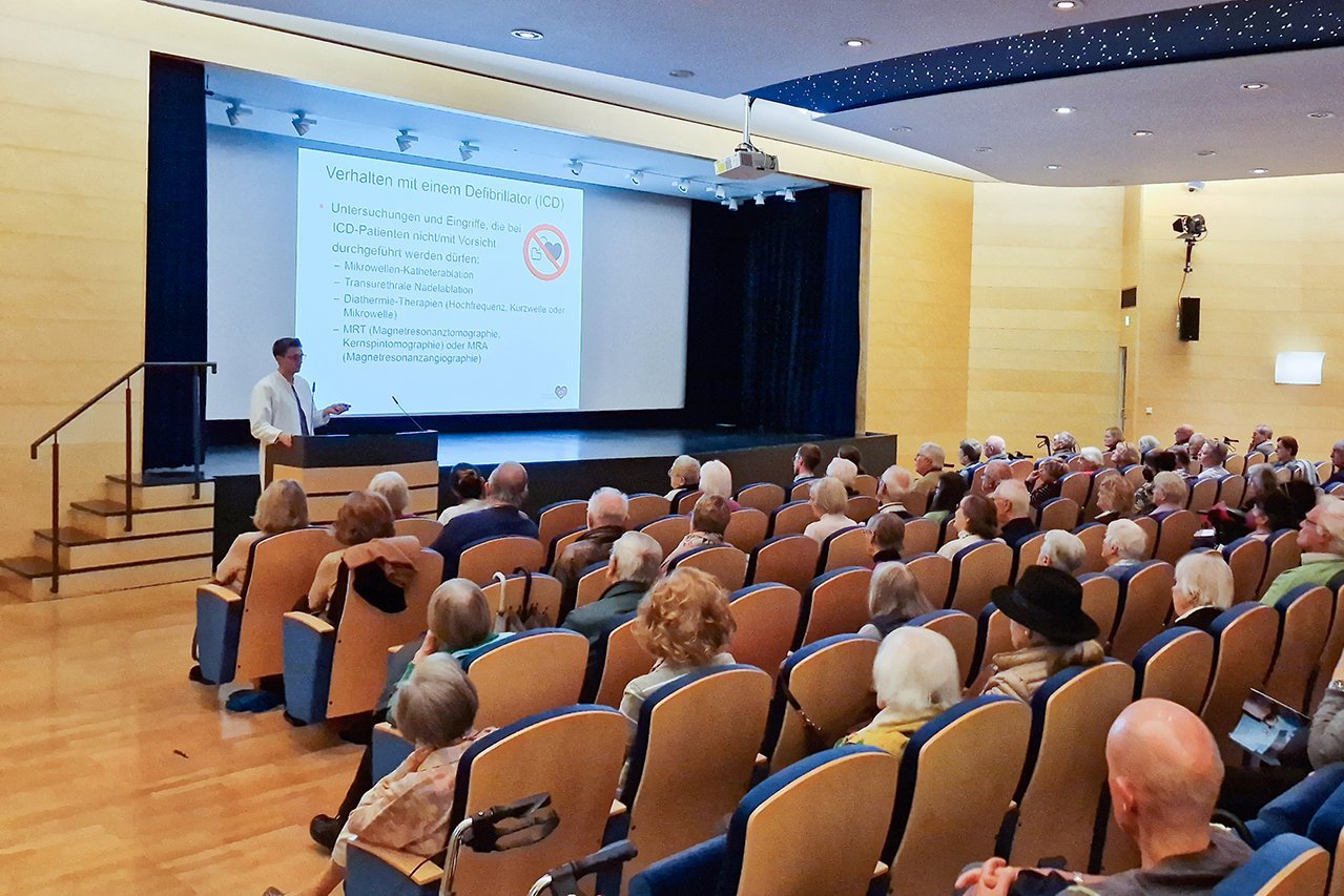 Augustinum Klinik Herzwochen Dr. Renz Besucher*innen sitzen im Theatersaal während Dr. Renz einen Vortrag hält