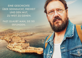 Bärtiger Mann mit Brille blickt geradeaus hinter ihm die Umrisse einer Stadt am Meer Bärtiger Mann mit Brille blickt geradeaus hinter ihm die Umrisse einer Stadt am Meer