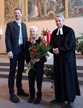 Augustinum-Geschäftsführer Tobias Geiger, Pfarrer Christian Gerstner und - in der Mitte - die Seniorin Elfriede Patz, die einen Blumenstrauß in den Händen hält  
