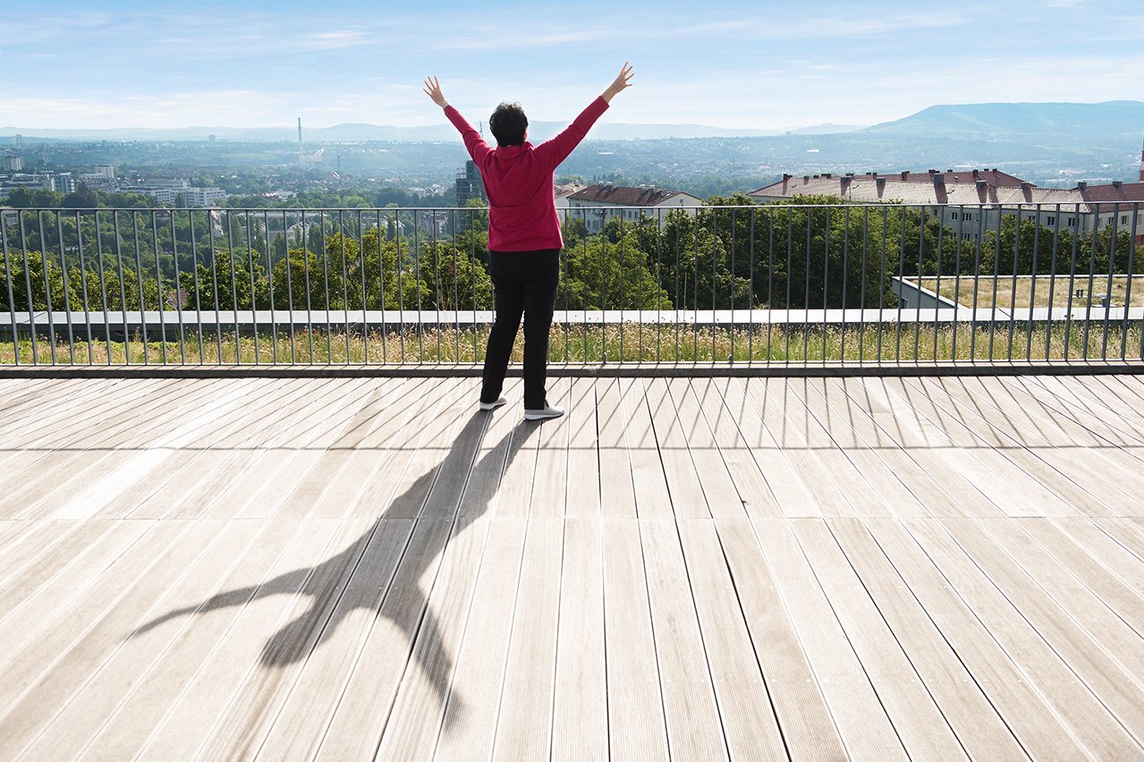 Augustinum Seniorenresidenzen Qi Gong Ein Seniorin steht mit nach oben gestreckten Armen auf einer Terrasse