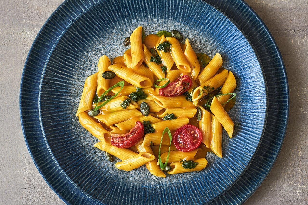 Auf einem tiefen blauen Teller mit breiterem blauem Unterteller ist breitgefächert Pasta angerichtet. Man erkennt getrocknete Tomaten, Pesto und und Kürbiskerne.