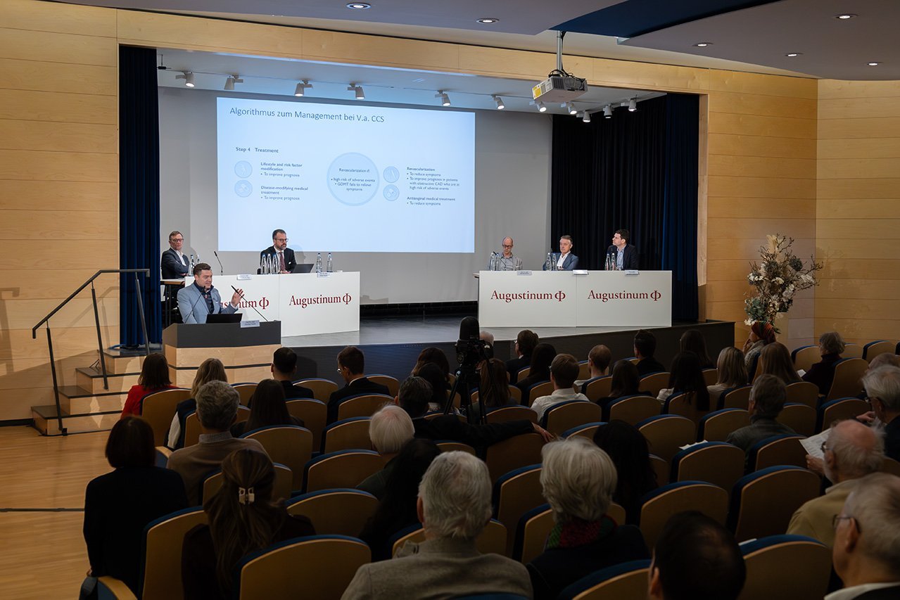 Augustinum Klinik Symposium Tesche Im Vordergrund ist ein Teil des Publikums, das auf den referierenden Oberarzt Dr. Tesche und die dahinterliegende Bühne blickt