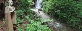 Skulptur der Regentrude vor einem Wasserfall mit Bachlauf