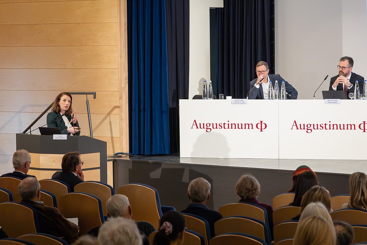 Augustinum Klinik Symposium Buiatti Eine Oberärztin der Augustinum Klinik, Dr. Buiatti, spricht zum Publikum, im Hintergrund sitzen zwei Veranstalter auf der Bühne