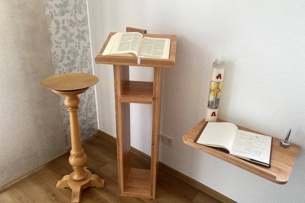 Eine Kerze und ein Notizheft auf einem kleinen Holztisch neben einem aufgeschlagenen Buch auf einer Holzablage