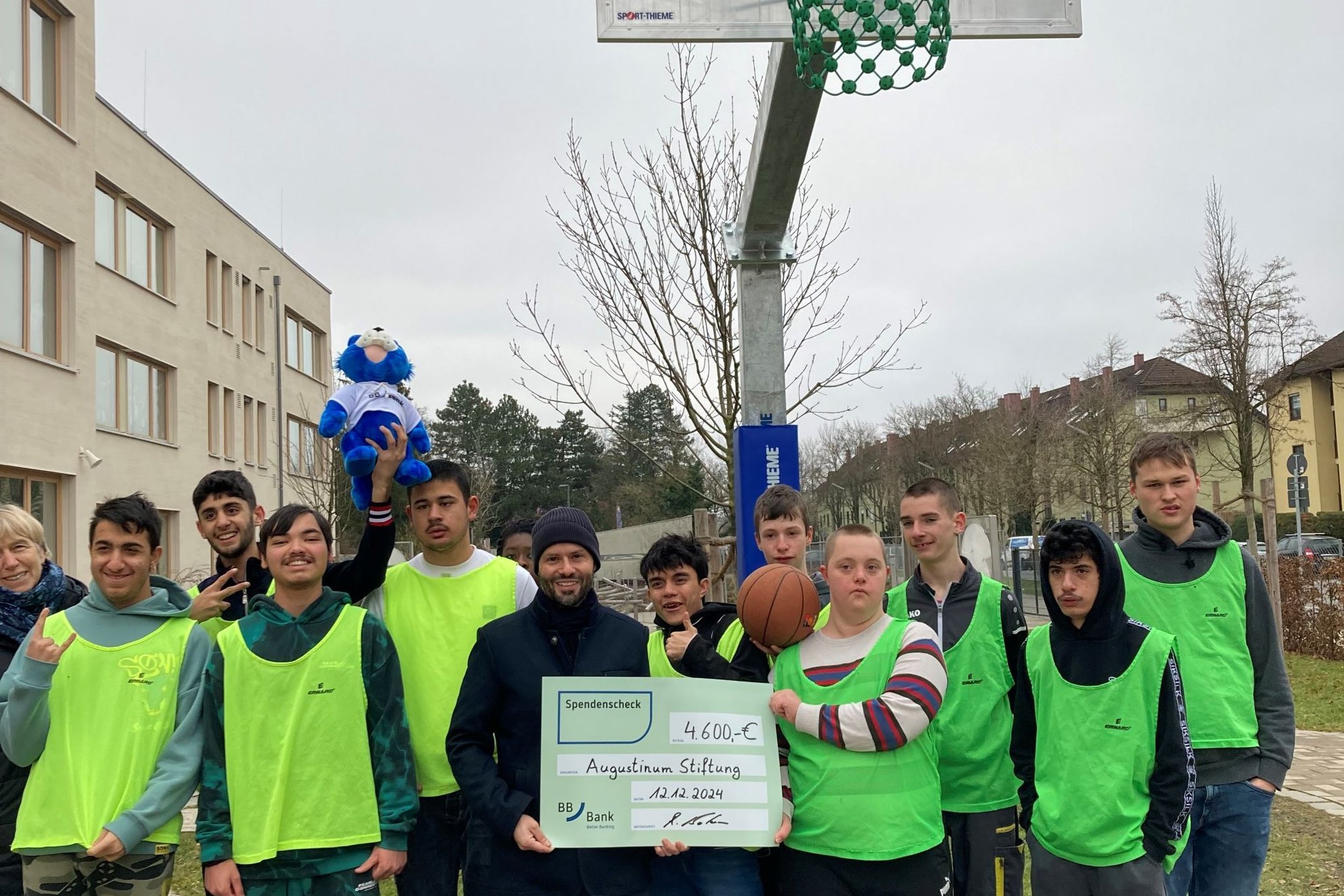 Augustinum Otto-Steiner-Schule Basketballkorb Schüler*innen vor Basketballkorb und Mann von BBBank, der Spendenscheck in den Händen hält, stehen vor einem Basketballkorb im Pausenhof