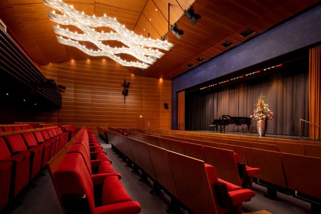 Bis zu 250 Zuschauer*innen können im Theatersaal Konzerte, Vorträge oder Filmvorführungen verfolgen. Ein Theatersaal mit roten Sitzen und einer Bühne, auf der ein Konzertflügel steht.