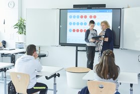 Eine Lehrerin und ein Schüler stehen vor der Klasse, hinter ihnen befindet sich ein Whiteboard