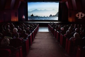 Augustinum Heidelberg WunderWelten Besucher*innen sitzen in einem dunklen Theatersaal und schauen auf die Leinwand, auf der ein ein Gewässer mit Bergen im Hintergrund zu sehen ist