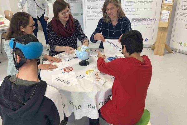 Augustinum Otto-Steiner-Schule Ausstellung Weltreligionen Drei Erwachsene und zwei Schüler sitzen gemeinsam an einem Tisch und beschäftigen sich mit den Weltreligionen