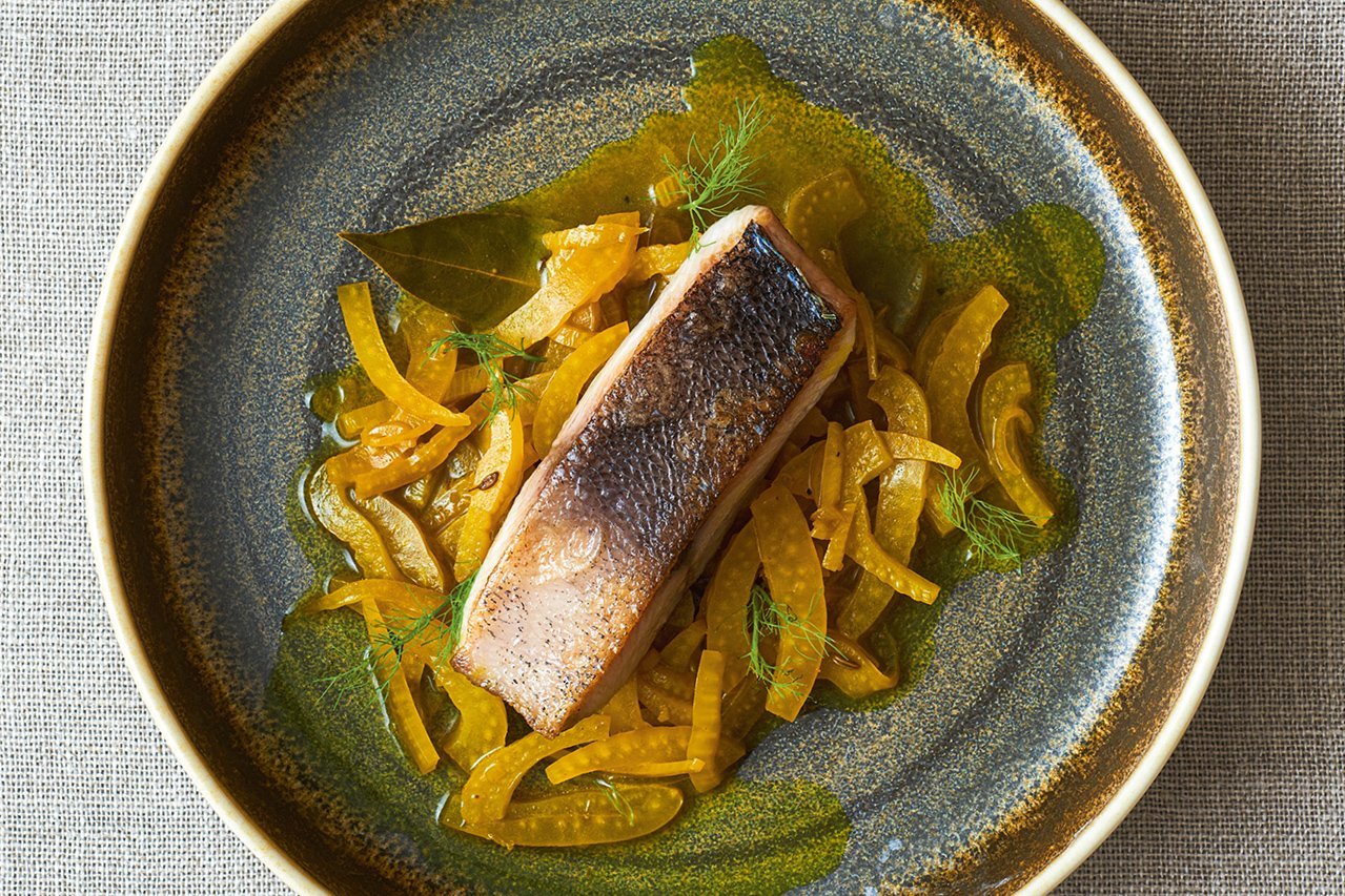 Das Bild ist gold-grau gehalten. In der Mitte einer tiefen runden brauen-grauen Tellers ist gelber Fenchelsalat angerichtet. Darauf liegt ein längliches Stück gebratener Fisch.