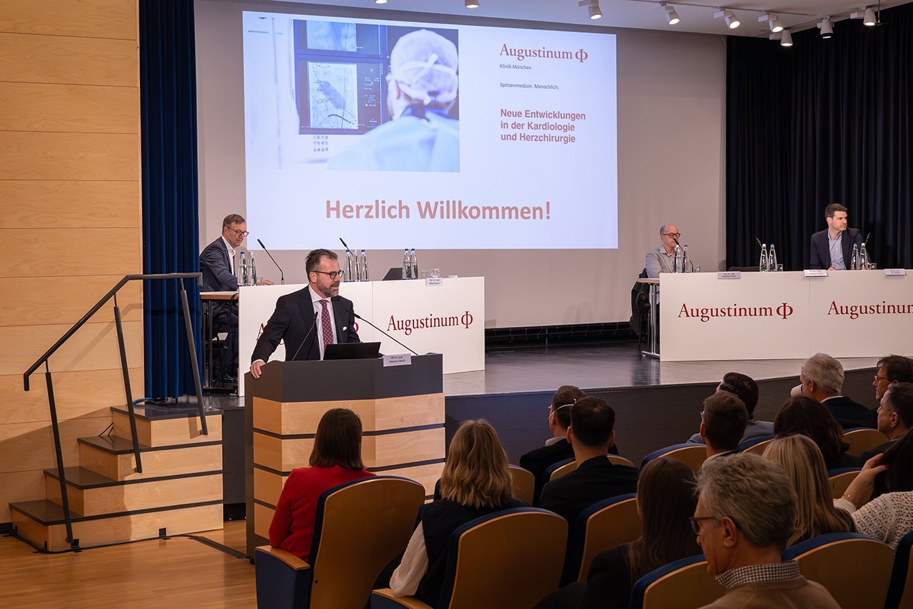 Augustinum Klinik Symposium Begrüßung Dr. Husser, ein Chefarzt der Augustinum Klinik, steht an einem Rednerpult und spricht zum Publikum. Weitere Veranstalter sitzen auf der Bühne