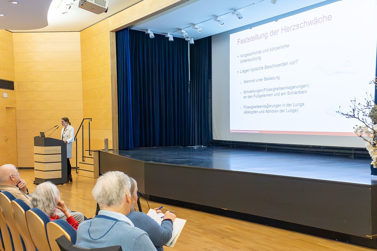 Augustinum Klinik Herzwochen von Jeinsen Besucher*innen betrachten die Präsentation im Theatersaal während eine Dozentin am Rednerpult einen Vortrag hält