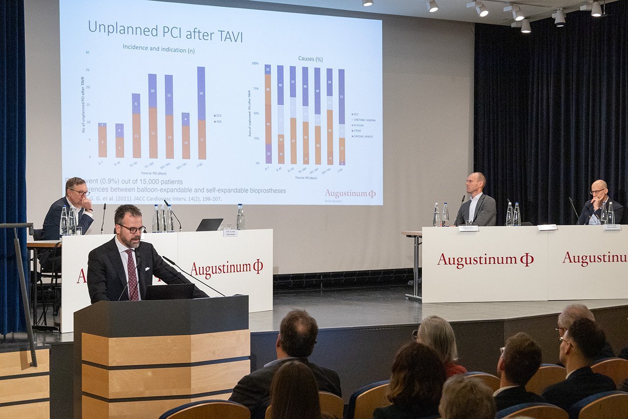 Augustinum Klinik Symposium Husser Dr. Husser steht vor der Bühne am Rednerpult und spricht in das Mikrofon, im Hintergrund ist seine Präsentation auf der Leinwand zu sehen und das Veranstaltungsteam sitzt auf der Bühne