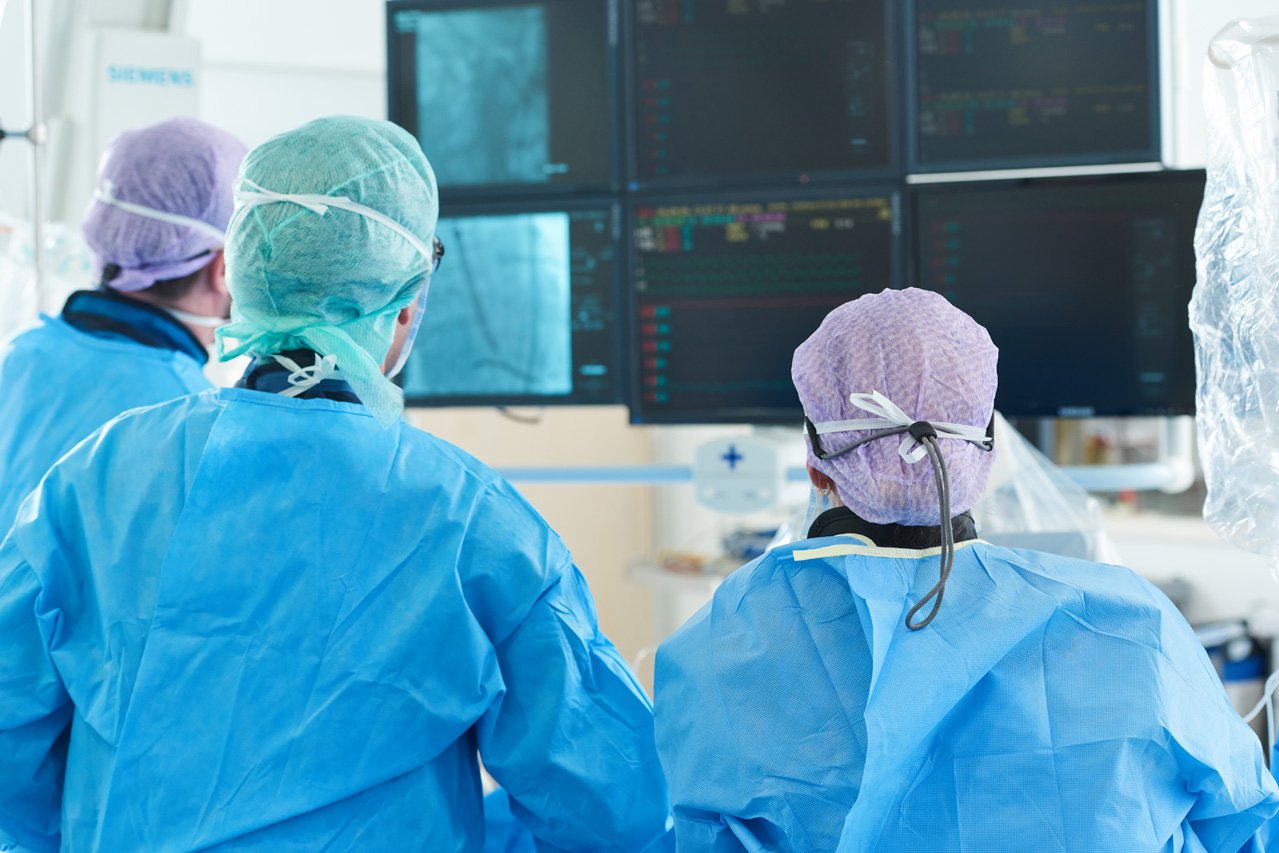 TAVI Drei Ärzte vor mehreren Monitoren kontrollieren den Fortschritt beim Einsetzen einer Aortenklappenprothese