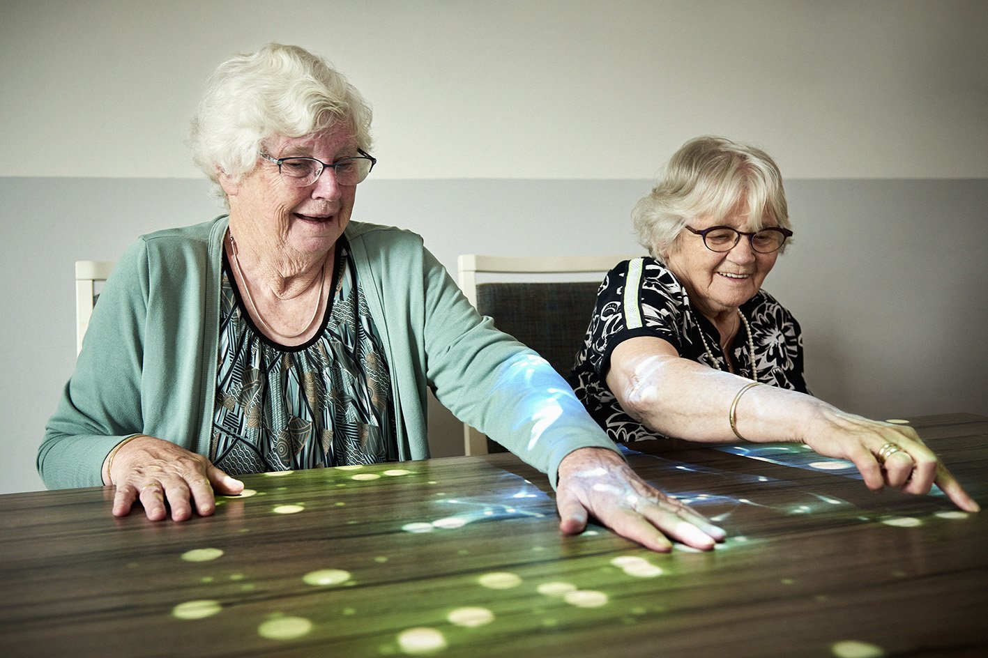 Augustinum Magazin Tovertafel Zwei Seniorinnen sitzen am Tisch und spielen ein Spiel, das mithilfe einer Tovertafel auf den Tisch projiziert wurde