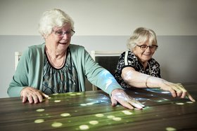 Zwei Seniorinnen sitzen am Tisch und spielen ein Spiel, das mithilfe einer Tovertafel auf den Tisch projiziert wurde
