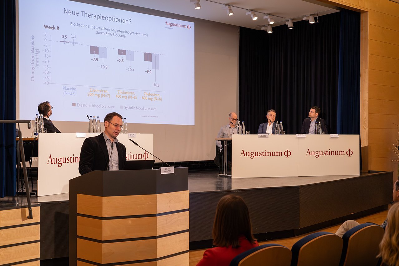 Augustinum Klinik Symposium Patzelt Der Dozent Dr. Patzelt steht vor der Bühne an einem Rednerpult und im Hintergrund sitzt das Veranstaltungsteam auf der Bühne