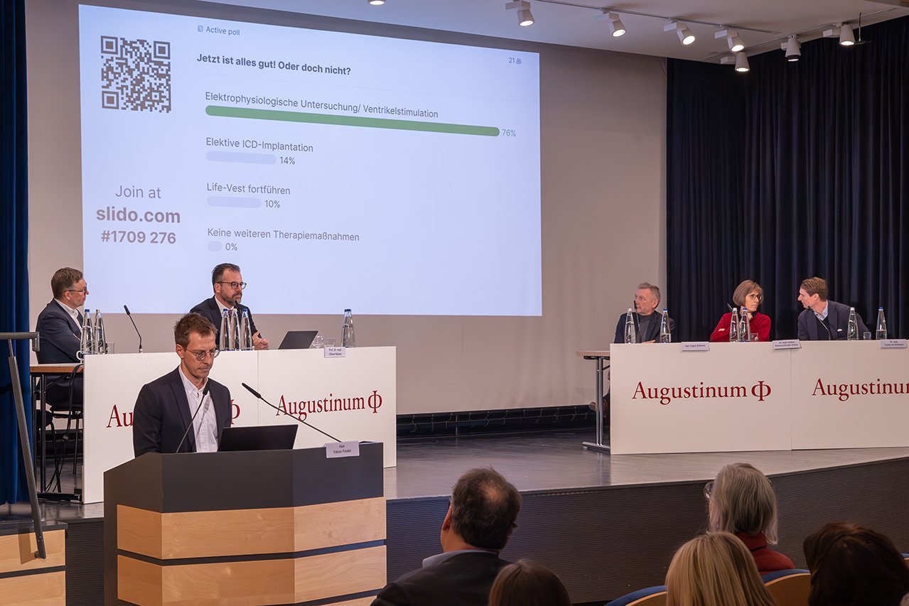 Augustinum Klinik Symposium Friedel Fabian Friedel referiert am Rednerpult während das Veranstaltungsteam im Hintergrund auf der Bühne sitzt und eine Umfrage auf die Leinwand projiziert wird