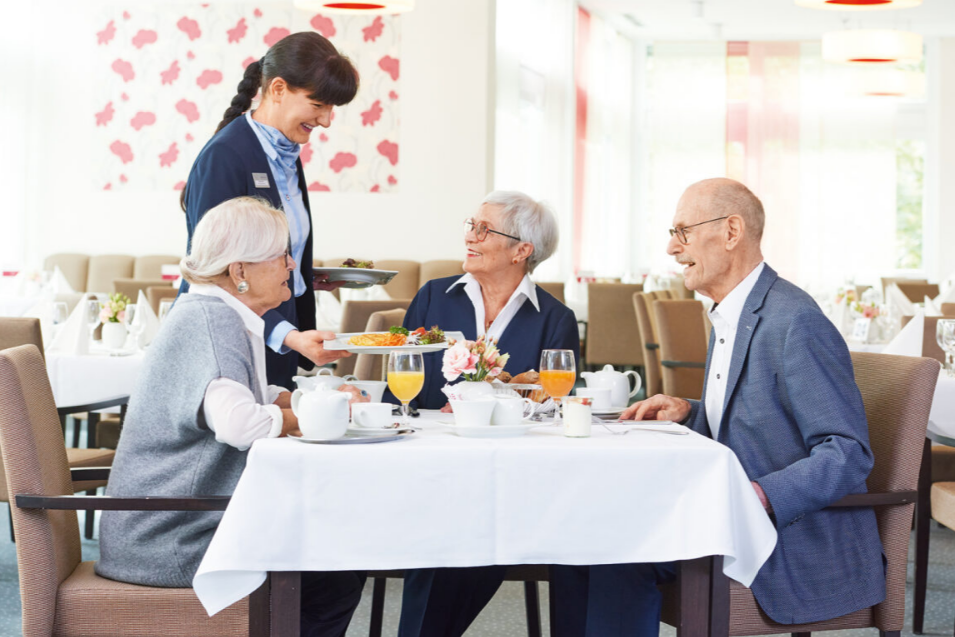 Drei Senior*innen werden beim Frühstück von einer Mitarbeiterin des Augustinum bedient