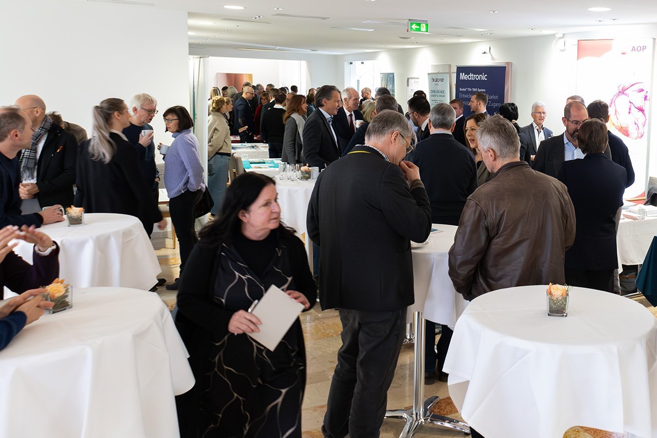 Augustinum Klinik Symposium Aussteller Viele Besucher*innen der Veranstaltung stehen in einem Raum mit kleinen weißen Tischen und unterhalten sich