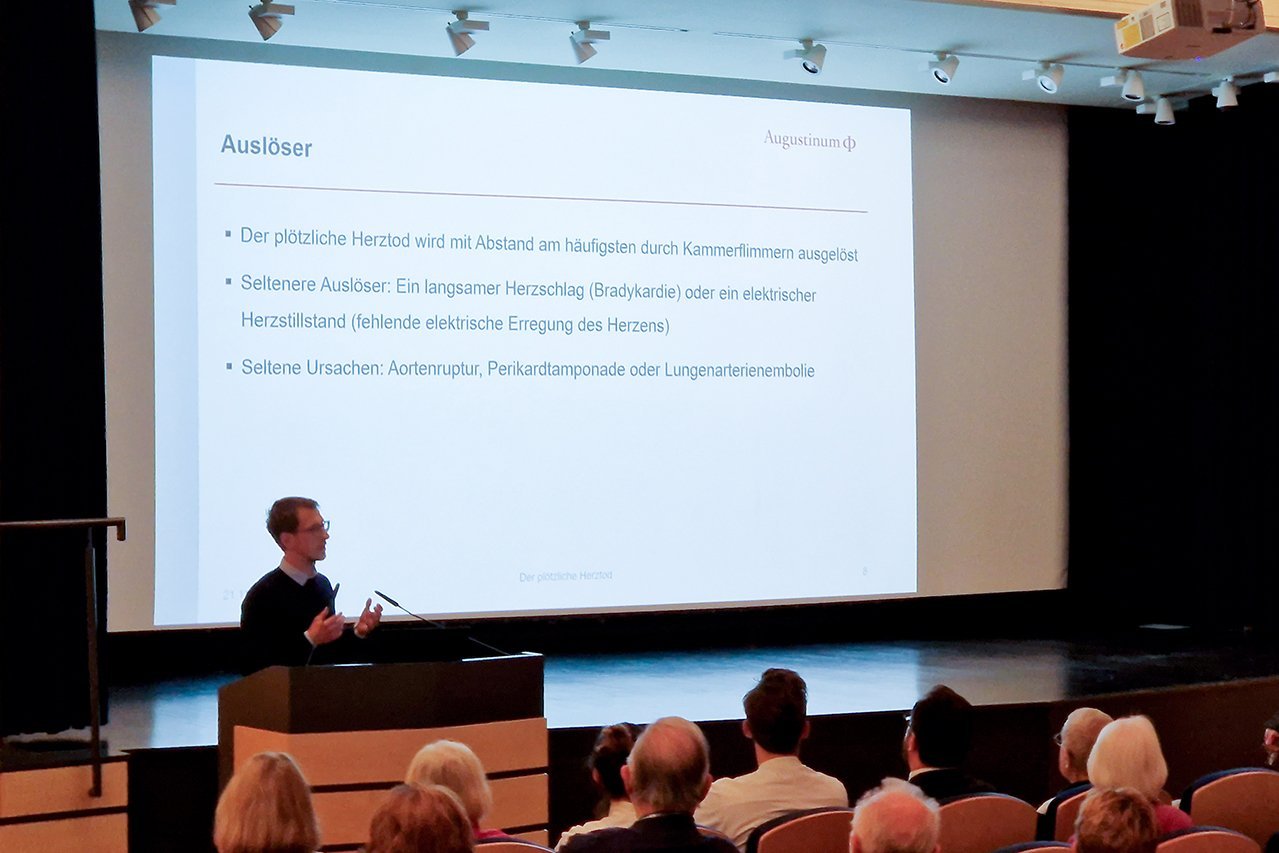 Augustinum Klinik Herzwochen Dr. Friedel Dr. Friedel hält einen Vortrag im besetzten Theatersaal, während seine Präsentation im Hintergrund auf eine Leinwand projiziert wird