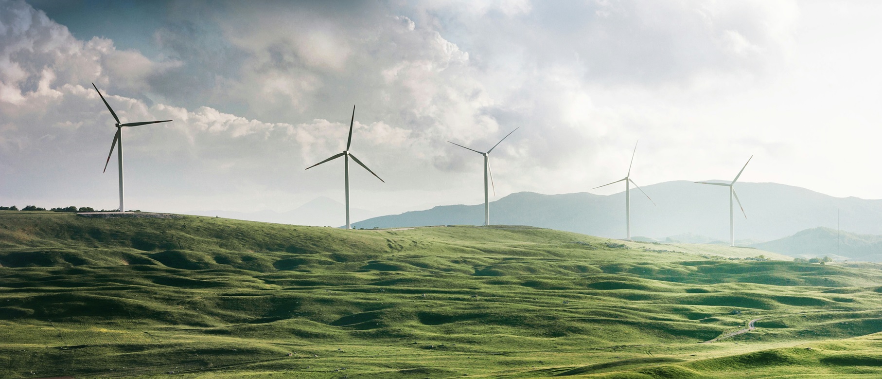 Augustinum Nachhaltigkeit Slider Fünf Windräder stehen auf einer hügeligen, grünen Ebene.