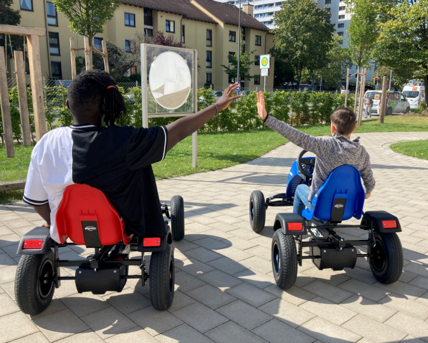Augustinum Fördermagazin Kettcars Eine Aufnahme von hinten, bei der zwei Schüler auf Kettcars sitzen und deren Arm zur jeweils anderen Person strecken